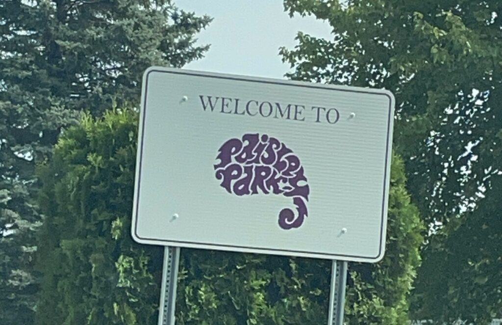 A square white Welcome To Paisley Park sign with letters in purple, in the shape of a paisley pattern.