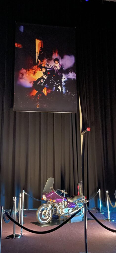 Purple rain poster of Prince above the actual Purple Rain motorcycle below on a stage.